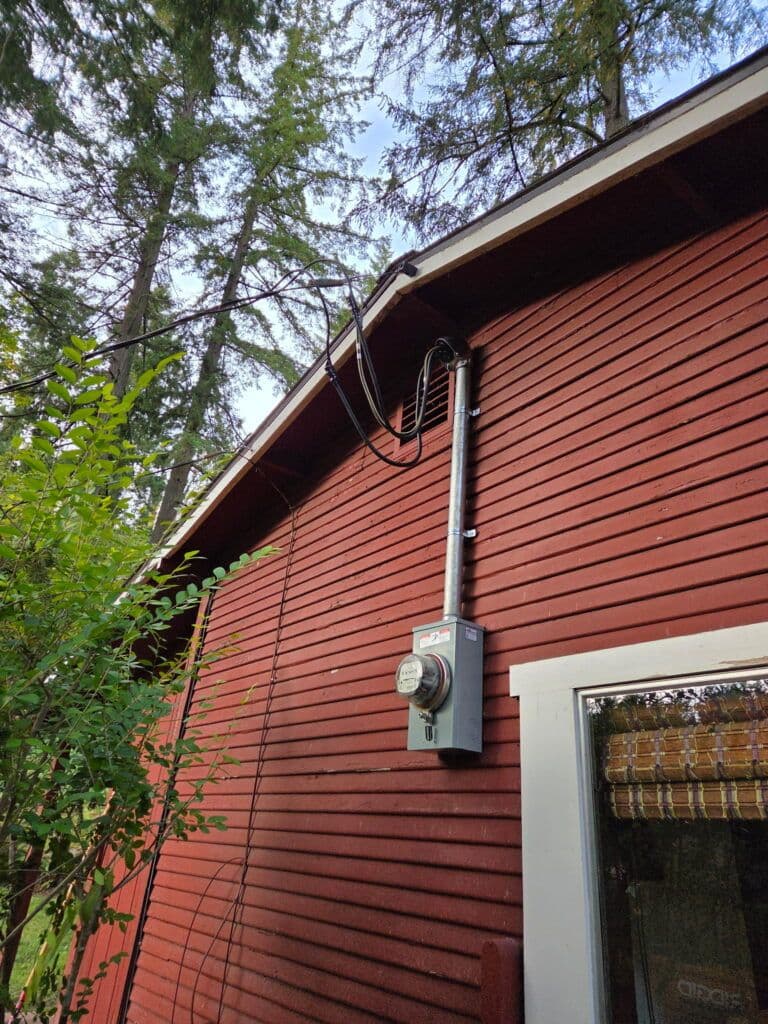 Electric meter installation on a red wooden house with trees and blue sky in the background.