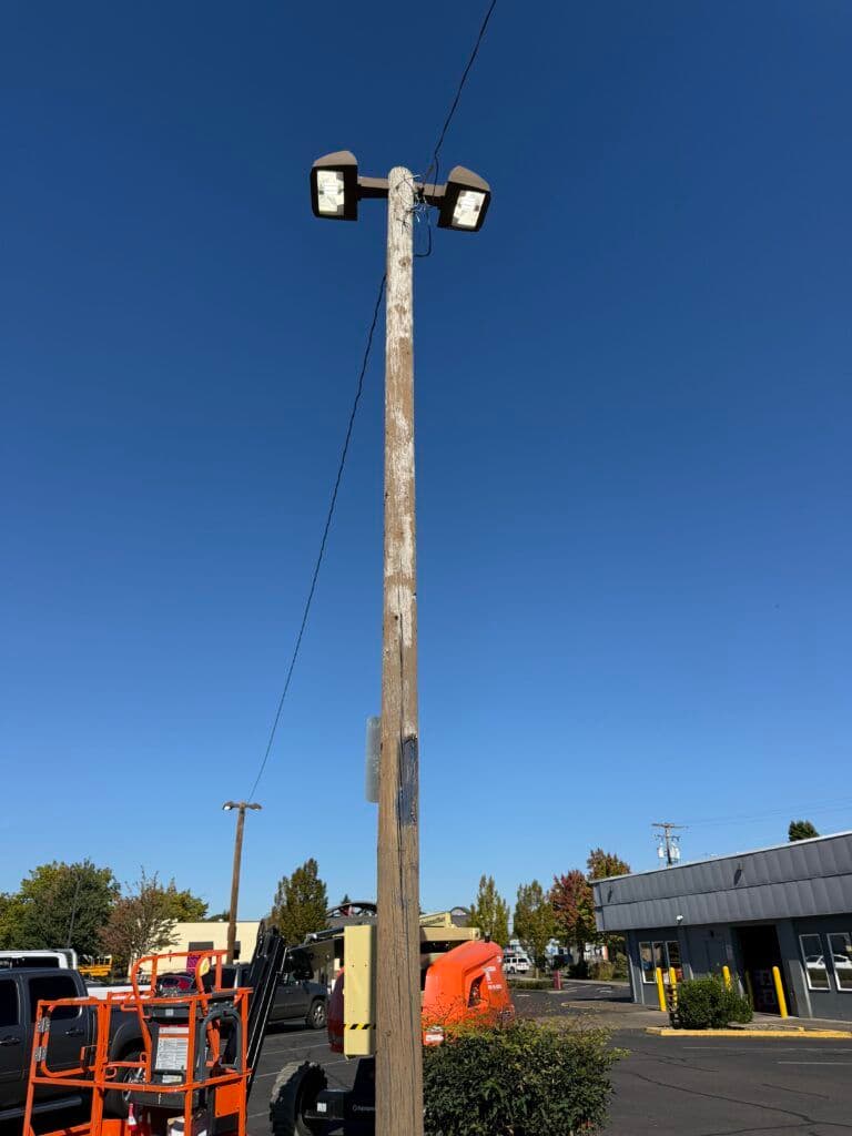 Wooden light pole with two floodlights against a clear blue sky in a commercial area.