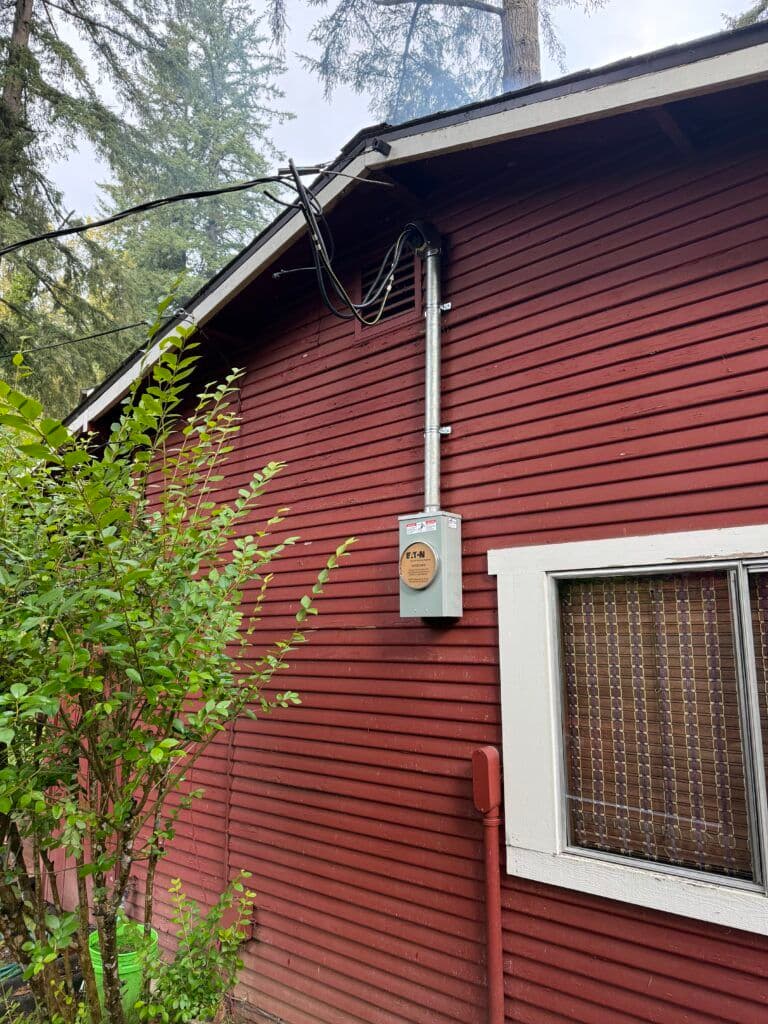 Electrical meter and wiring on the side of a red wooden house surrounded by greenery.